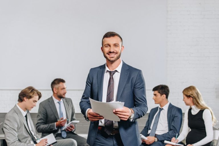 happy-business-coach-standing-in-front-of-businesspeople-at-training-in-hub.jpg